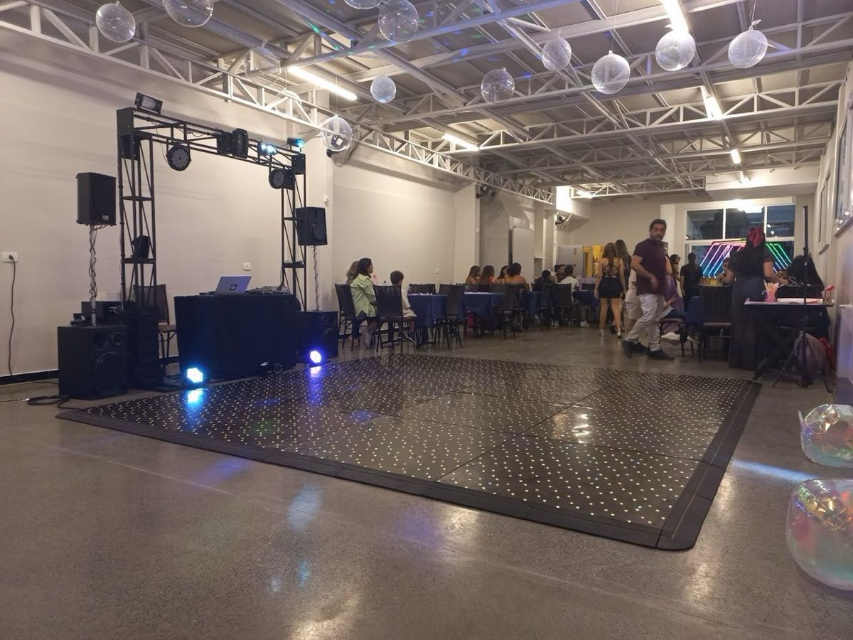 Booth do DJ Alexandre montado com truss, caixas de som e iluminação, com pista de LED e convidados ao fundo em salão de festas