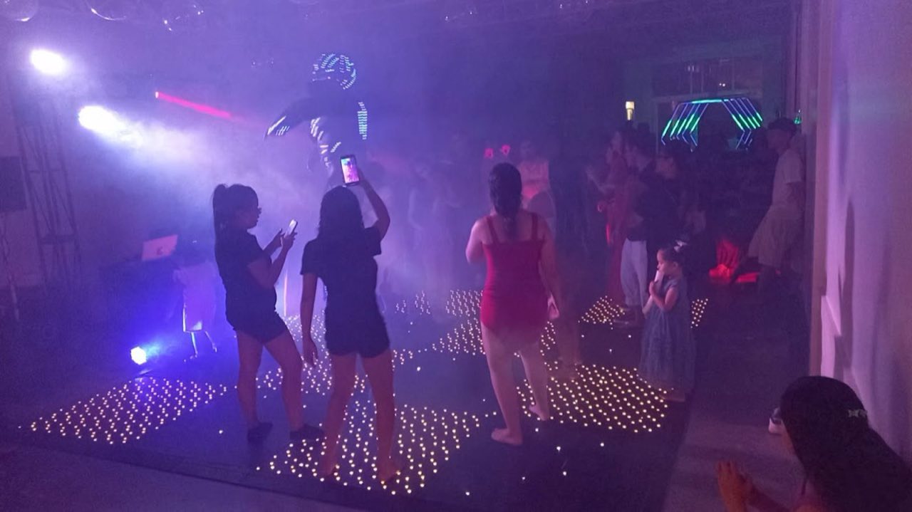 Pista cheia com convidados dançando e performer com traje de LED, efeito de fumaça